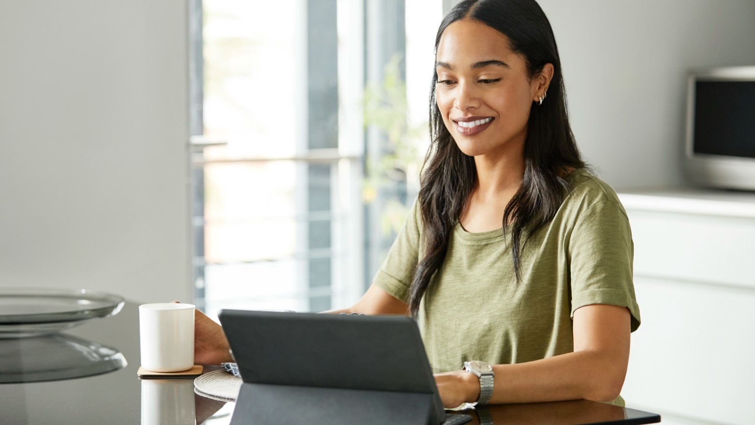 A woman sits at her kitchen counter with a mug and her tablet