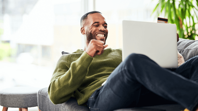 A man sitting on a couch, smiling and looking at this laptop while holding his credit card