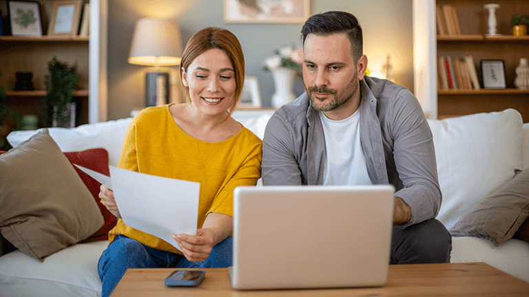 A couple sitting on a couch using a laptop and reviewing paperwork