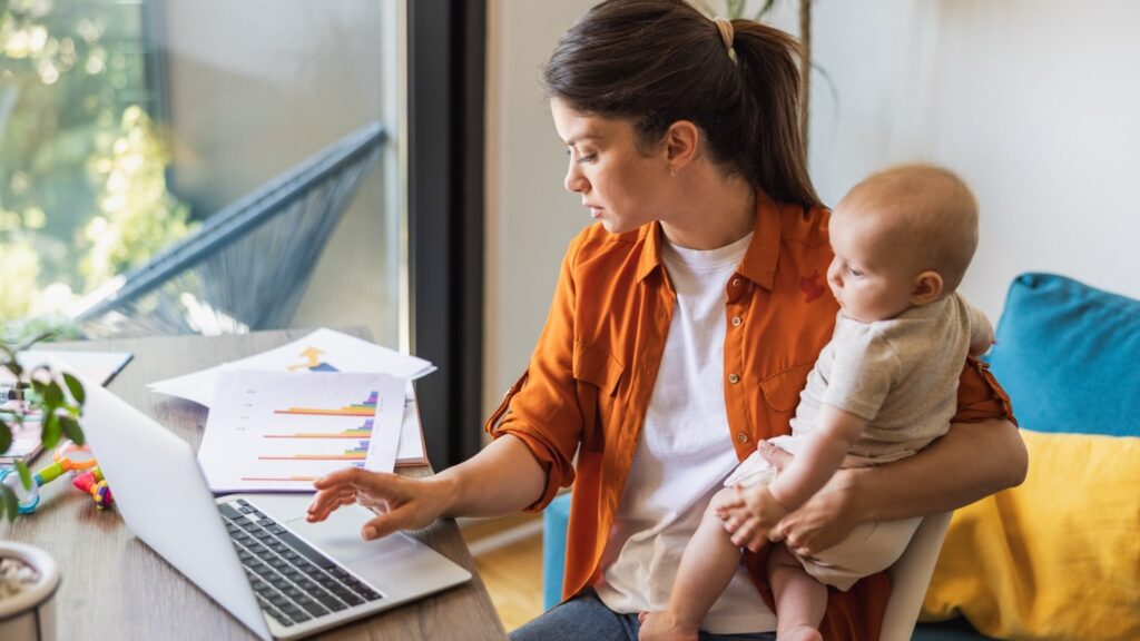 A woman holds her baby while working on her laptop